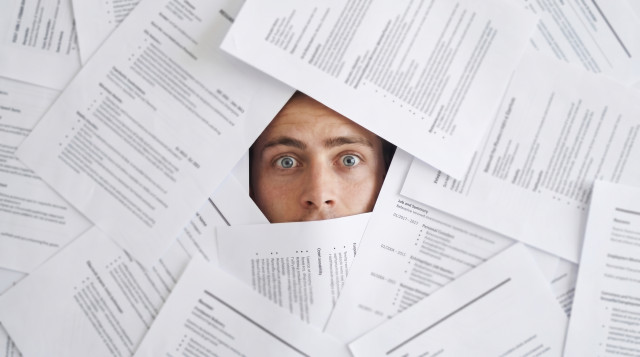 A man's panicked face barely visible among dozens of resumes and documents covering the entire frame, about to disappear under the pile.