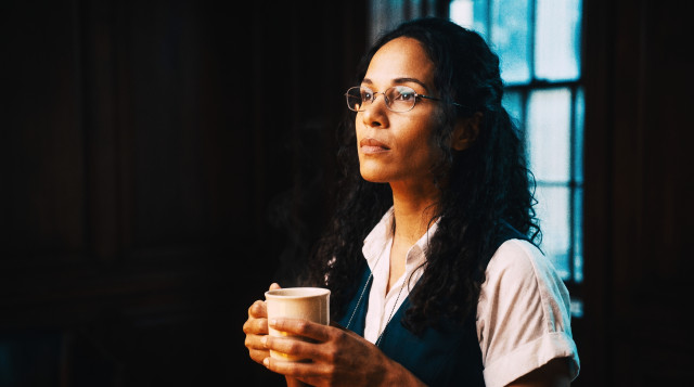 Thoughtful female recruiter with curly hair and glasses holding a cup of coffee in a dimly lit office at night