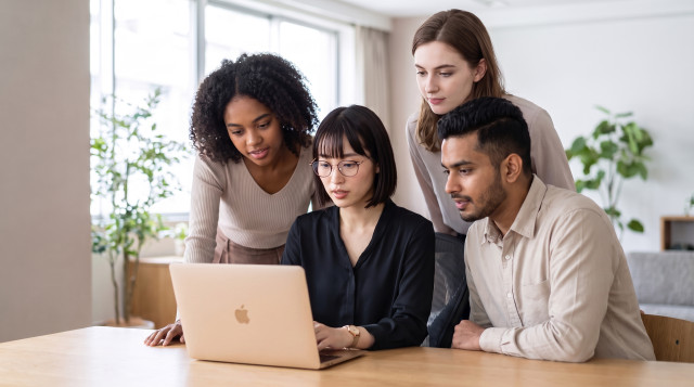 A diverse hiring team of four gathered around a laptop, reviewing candidates together in a bright office