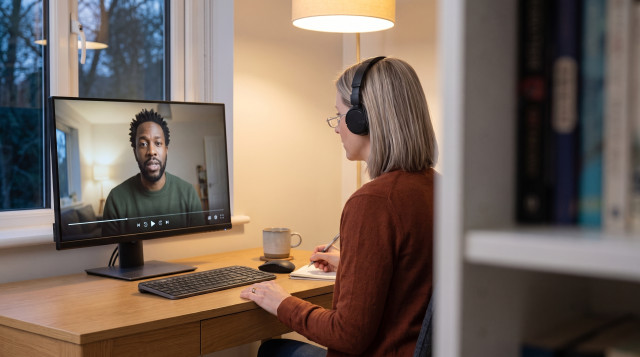 A recruiter wearing headphones takes notes at her home desk in the evening while watching a candidate's recorded video answer on her monitor.