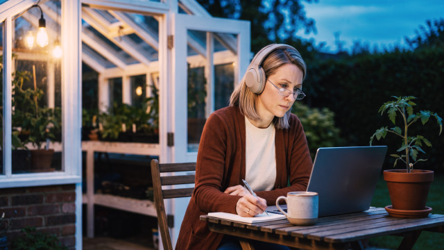 Recruiter in headphones reviewing one-way video interview answers on a laptop at a garden table by a greenhouse