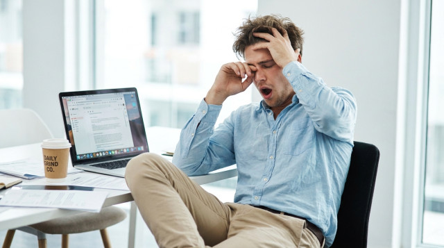 Tired startup founder in a blue shirt sitting at his desk, rubbing his eyes in exhaustion, surrounded by a laptop full of emails and a coffee cup
