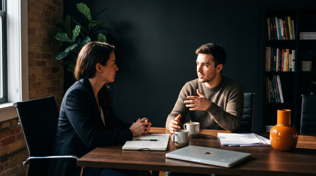 Interviewer and candidate in a structured job interview at an office table, candidate answering a follow-up question