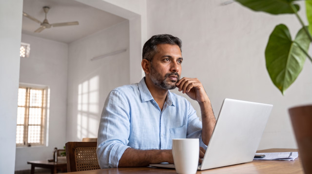 Man in a light blue shirt looking away from his laptop, thinking, coffee mug on the desk