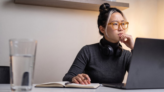 Woman in glasses and headphones frowning at a laptop, taking notes in a notebook